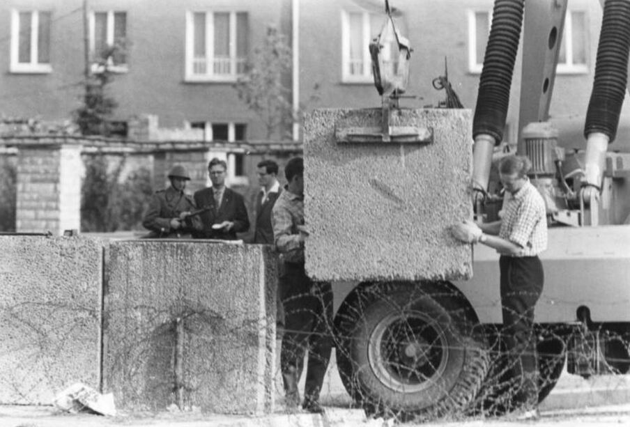 Kran lässt hinter Stacheldraht begleitet von Arbeiter Betonstück ab